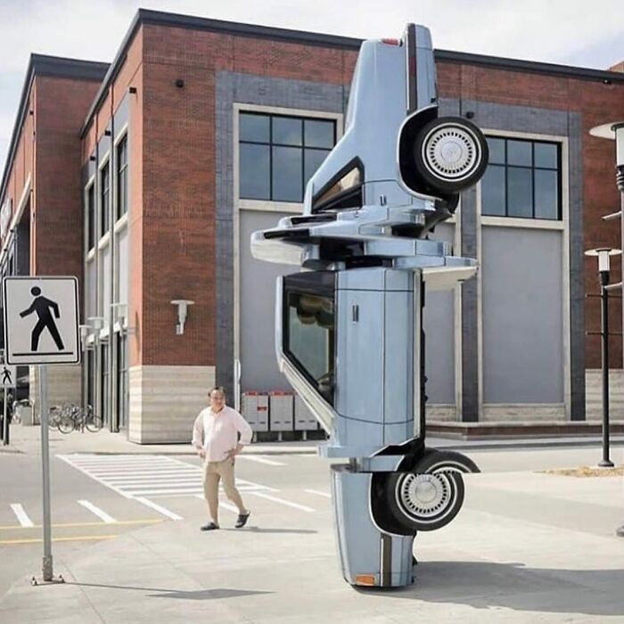 A unique street art installation featuring a light blue car standing vertically on the pavement in an urban setting.