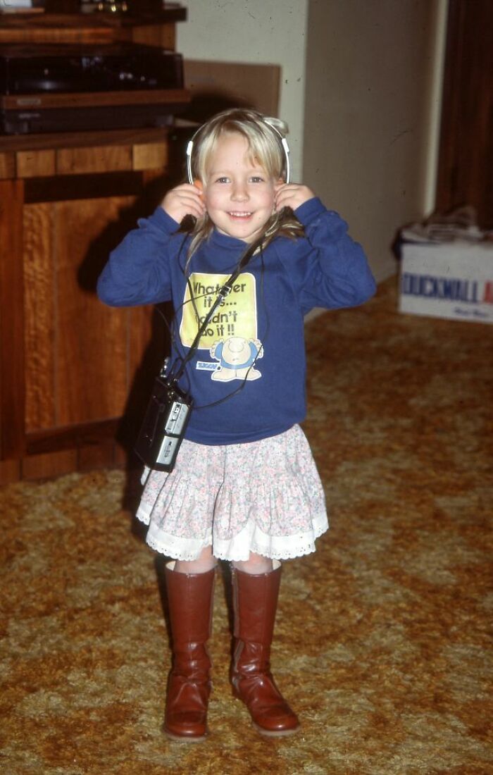 A young girl with blonde hair smiles while wearing retro tech headphones and holding a portable device, capturing the 80s weirdness.