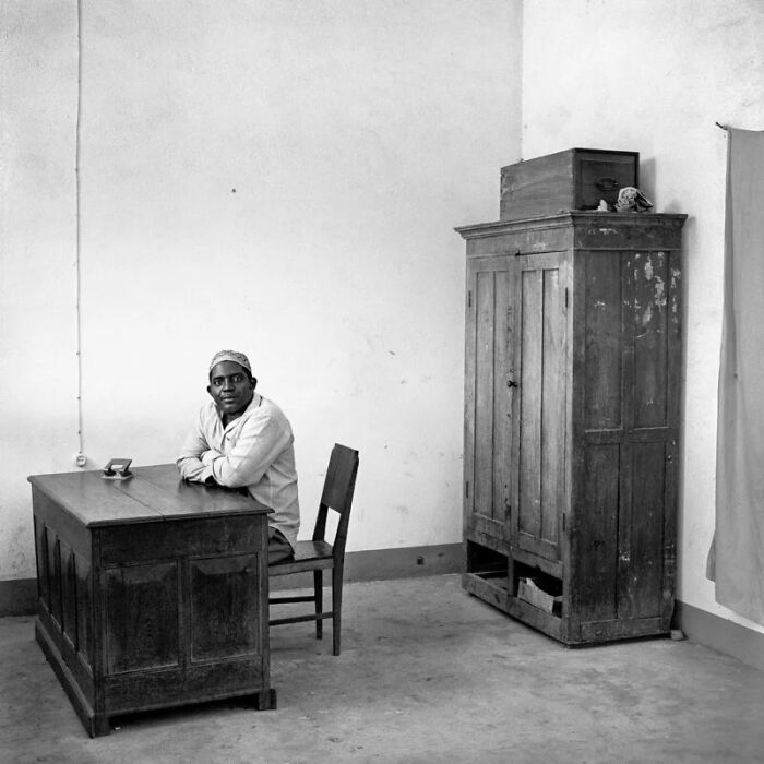 A man in a cap sits at a wooden desk in a minimalist office, documenting the people and offices behind bureaucracy.