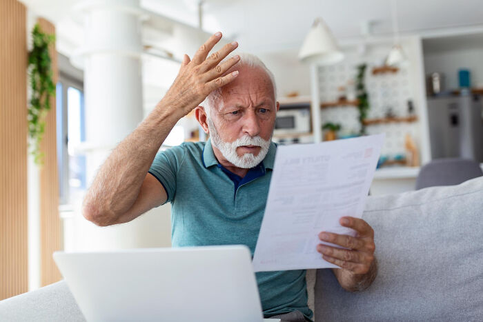 An elderly man looking confused at a document with a laptop, highlighting dumb ways people ruin their lives financially.