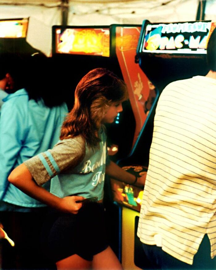 A person plays a retro tech arcade game, likely Pac-Man, in a dimly lit arcade, capturing the 80s weirdness.