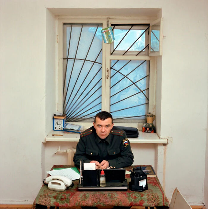 A man in a military uniform sits at a desk in front of a barred window, documenting the people and offices behind bureaucracy.