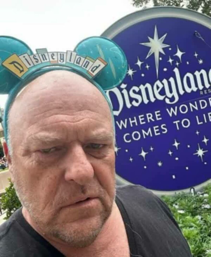 A man with a pained expression wearing Mickey Mouse ears in front of a Disneyland sign, a disturbing pic.