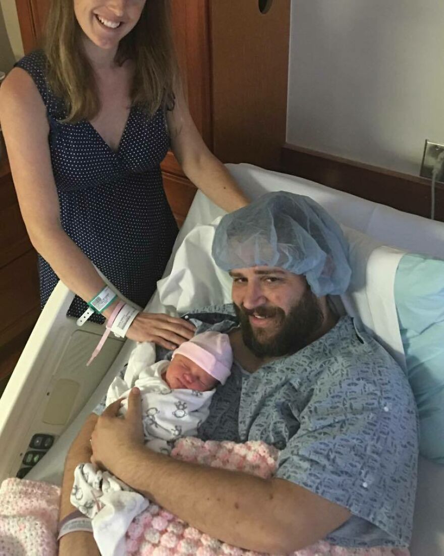 A man in a hospital gown holding a newborn baby with a woman smiling nearby, a heartwarming pic.