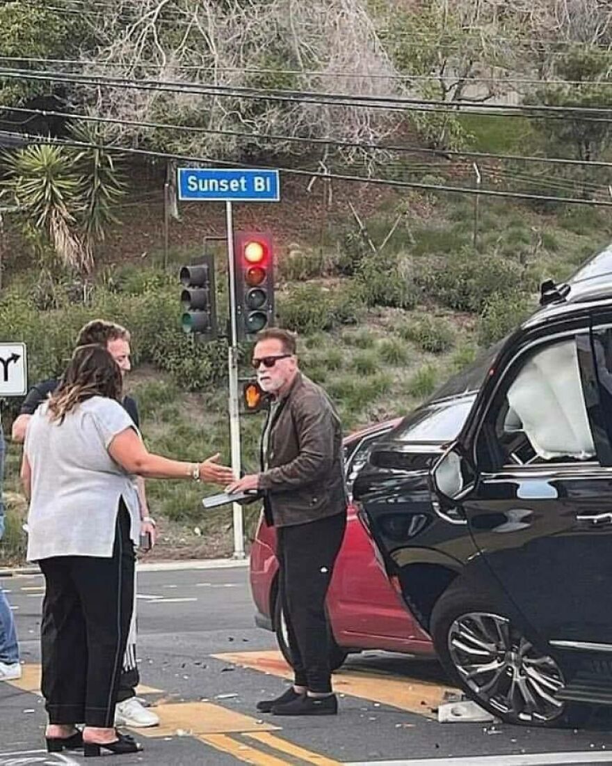 Arnold Schwarzenegger talking to a woman next to a damaged car on Sunset Blvd, a chaotic and uncomfortable scene.