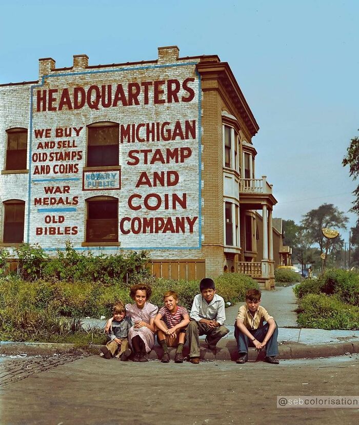 A historical colorized photo of children sitting on a curb in front of the Michigan Stamp and Coin Company.