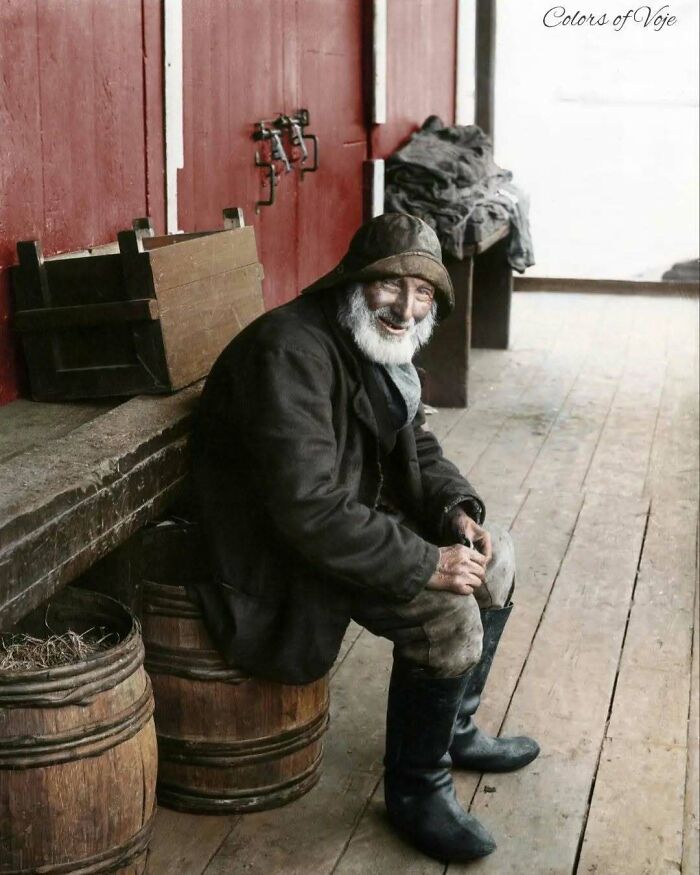 A historical colorized photo of an old man with a beard and a hat smiling while sitting on a wooden barrel. Stunning historical colorized photos.