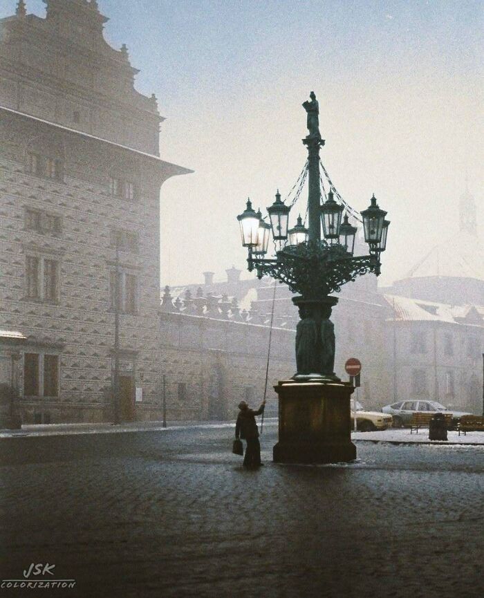 A person lighting a street lamp in a misty, historical colorized photo of a European city street.