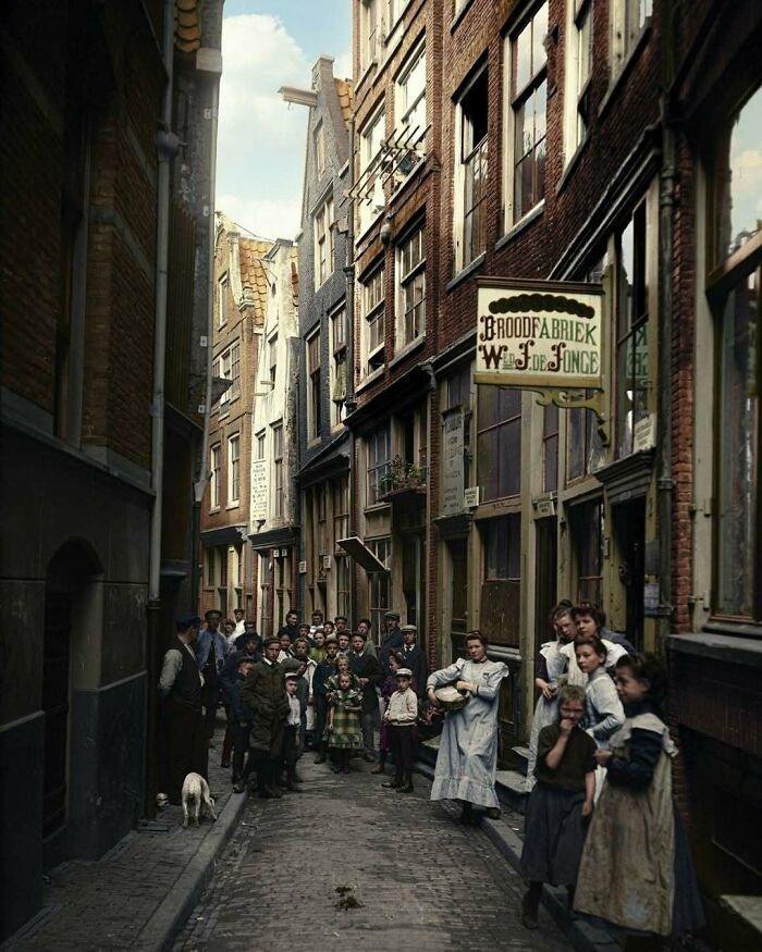 A narrow street with people and buildings, including a bakery sign. Stunning historical colorized photos.