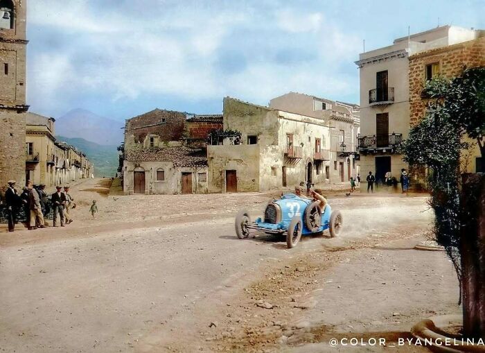 A historical colorized photo of an old racing car on a dirt road in a village, bringing the past days to life.