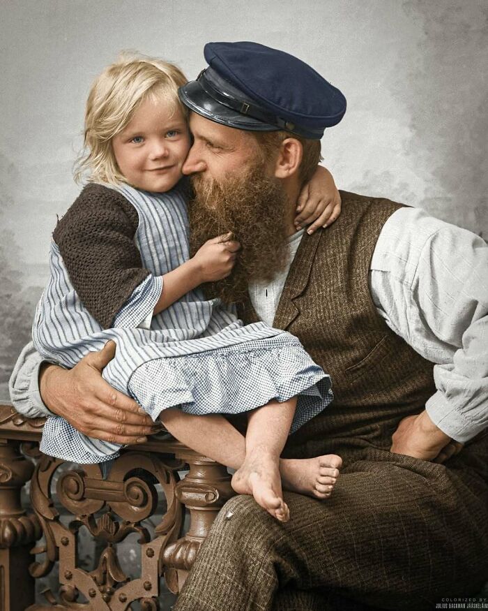 A stunning historical colorized photo of a bearded man in a captain's hat hugging a little girl.