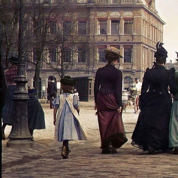 Historical colorized photo of women and a child walking on a street, with a historical building in the background.