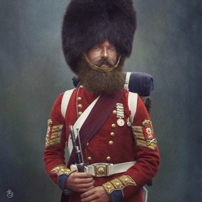 A historical colorized photo of a soldier in a red uniform and large fur hat, holding a rifle with a prominent beard.