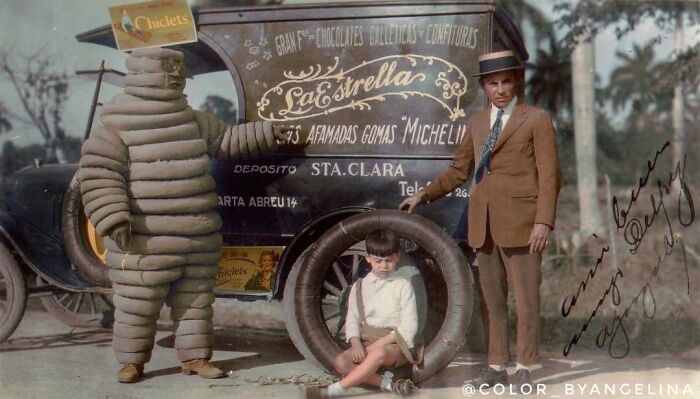 A Michelin Man mascot, a man in a suit, and a boy with a tire, next to a truck. Stunning historical colorized photos.