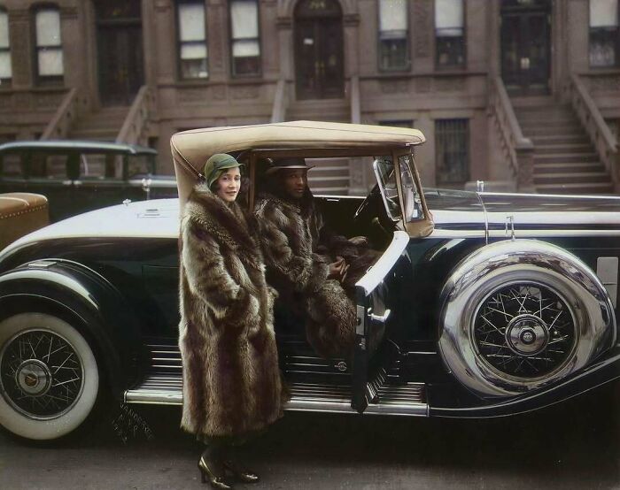 Historical colorized photos: Two elegantly dressed people in fur coats with a vintage car on a city street.