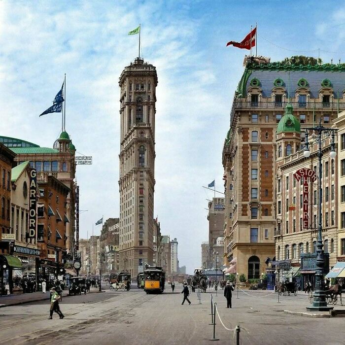 A vibrant historical colorized photo of a bustling city street with a tall building and streetcar under a blue sky.
