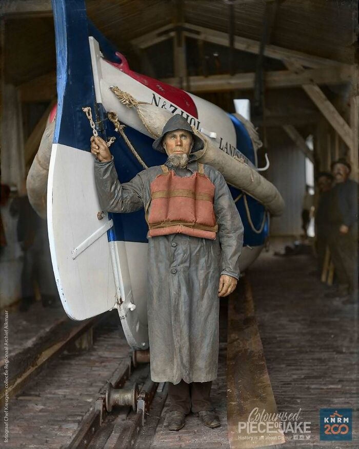 Historical colorized photo of a man in a rain jacket and hat, standing in front of a lifeboat in a boathouse.