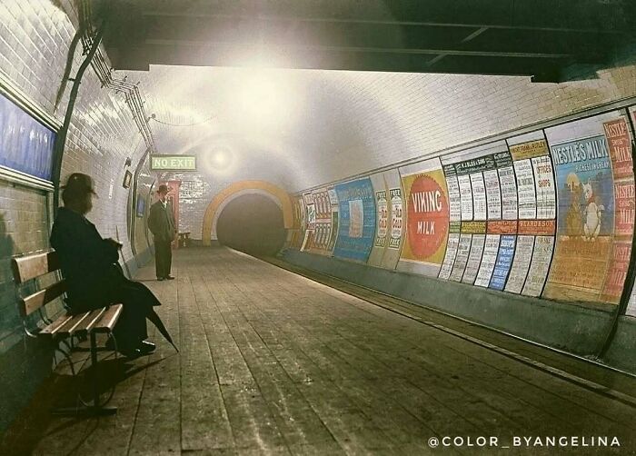 Historical colorized photo of an old subway station with people waiting on a bench and posters on the wall.