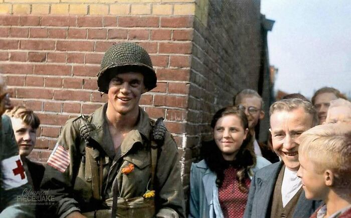A smiling soldier in a helmet, with an American flag patch, stands among a group of people in a historical colorized photo.