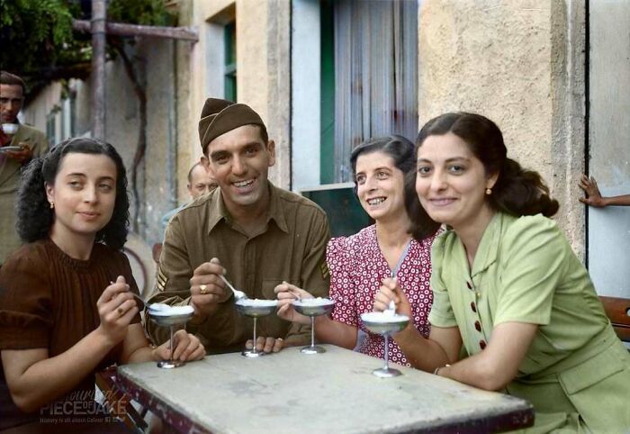 Historical colorized photo of four people, including a soldier, happily eating ice cream outdoors at a table.