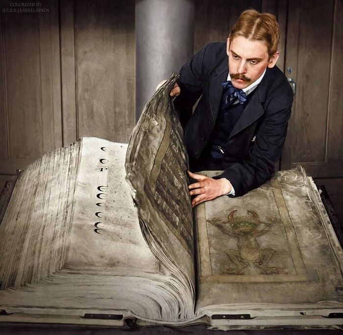 Historical colorized photo of a man in a suit and mustache looking at a huge, old, open book with a drawing.