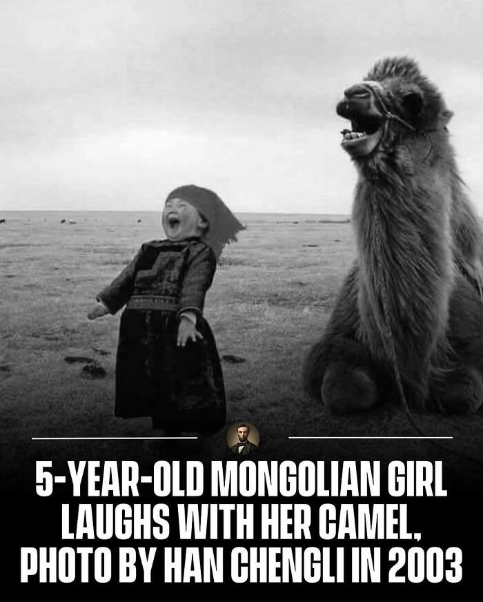 A photo from history of a young Mongolian girl laughing alongside a camel in a grassy field
