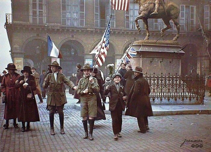 A historical colorized photo of people marching in a parade with flags and a statue in the background, making the past come to life.