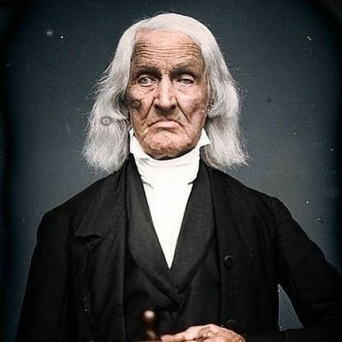 A stunning historical colorized photo of an elderly man with long white hair and a dark suit.