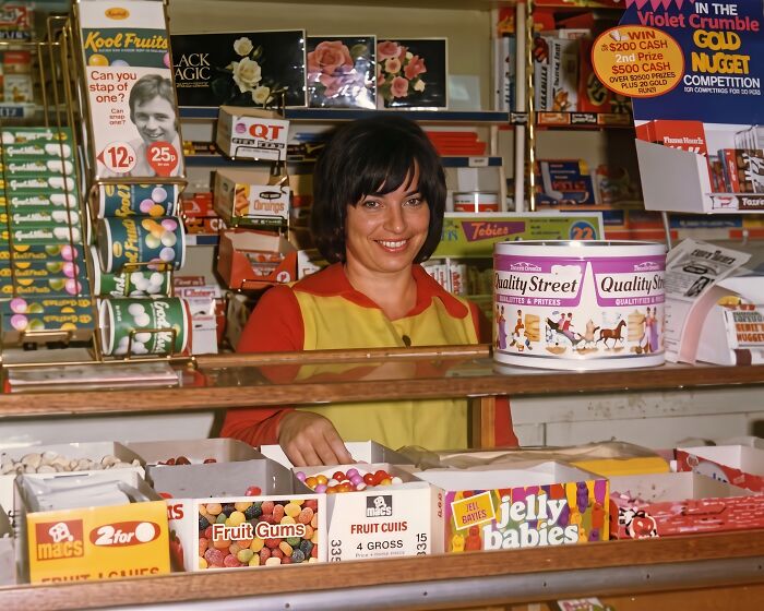 Smiling woman behind a counter in an old-fashioned shop filled with treats, bringing back Aussie memories.
