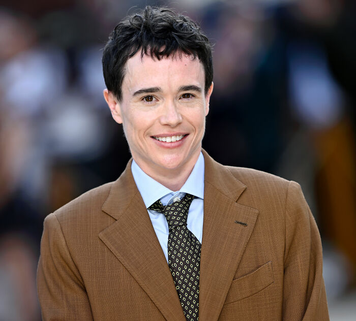 Trans actor Elliot Page smiling in a brown suit, exposing Hollywood's reality after 'The Odyssey' success. Trans actor Elliot Page smiling in a brown suit, exposing Hollywood's reality after 'The Odyssey' success.