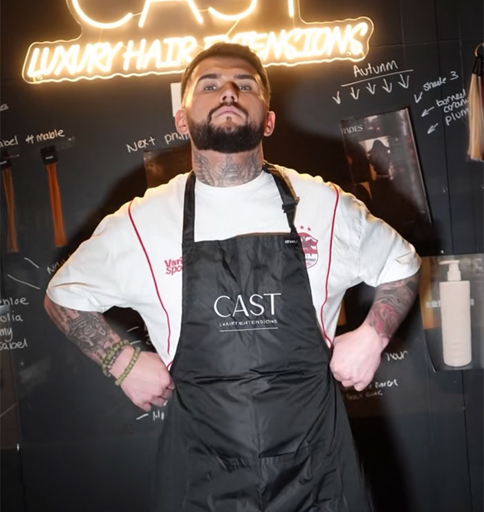 A top hairdresser in a black apron stands confidently in front of a salon wall, giving a brutal reality check.