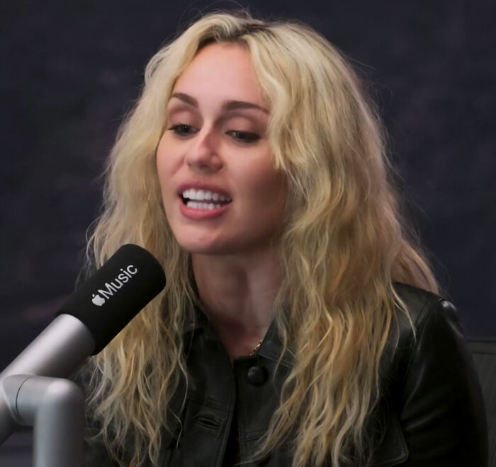 Miley Cyrus, with blonde wavy hair, speaks into an Apple Music microphone. She wears a black leather jacket.