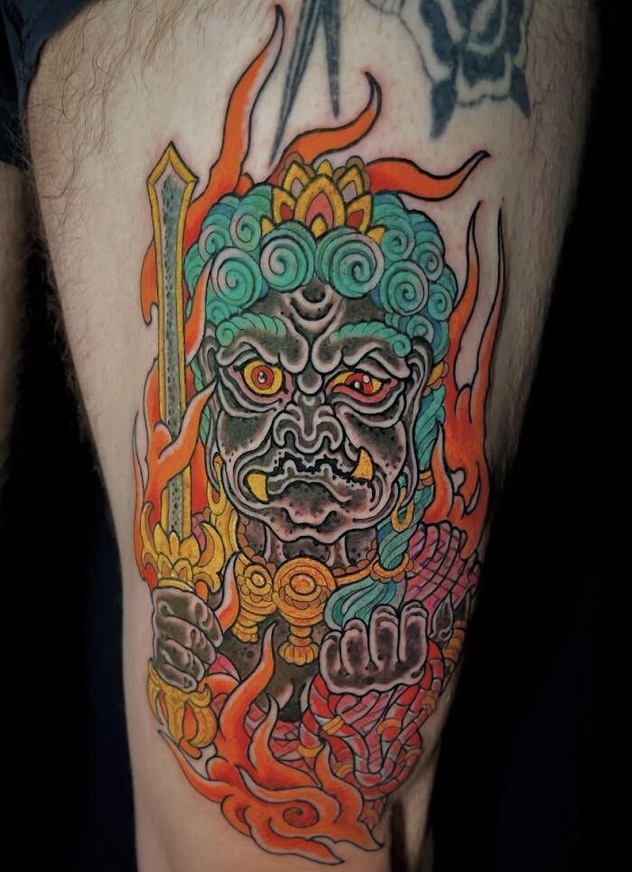 Stunning mythology tattoo of a colorful demon with a sword on a leg, from Sacred Gold Studio.