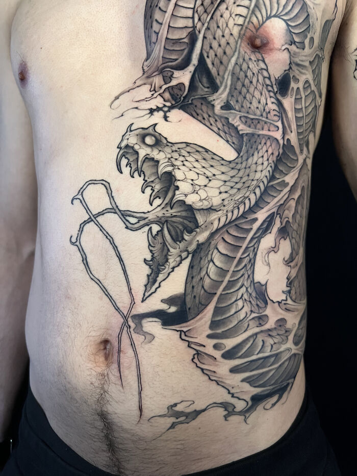 A large, detailed ancient mythology tattoo of a snake-like creature on a torso from Sacred Gold Studio.