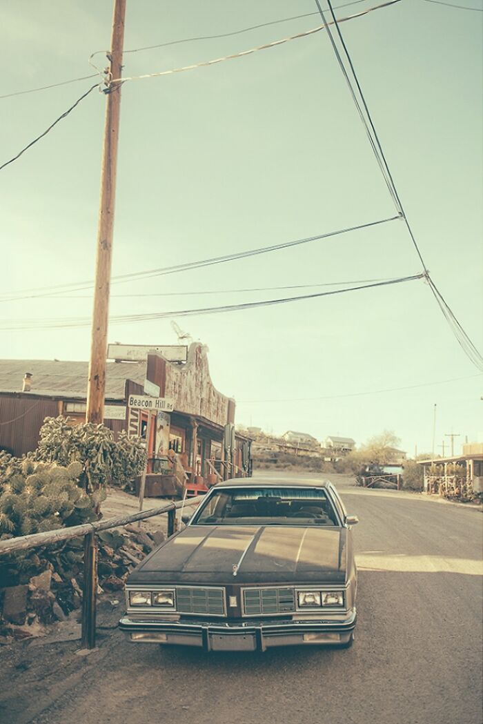 A classic car parked on a dusty street in a bygone American West town, featuring old buildings and utility poles.
