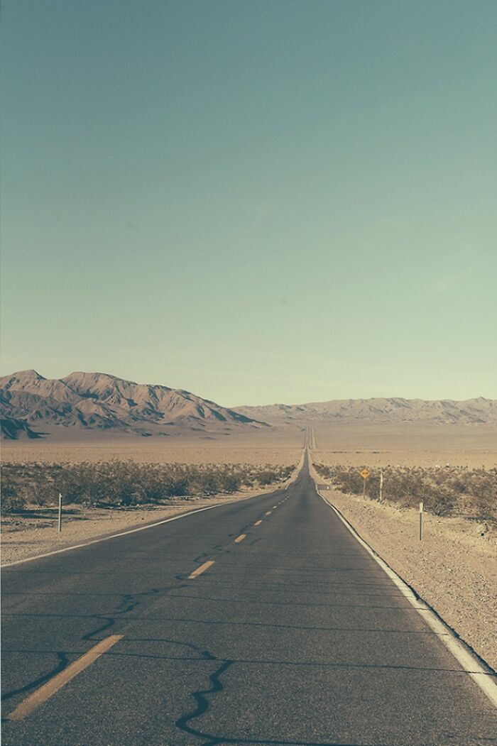 A long, straight road extends into the horizon through a desert landscape with mountains in the American West. Rusty and dusty remnants.