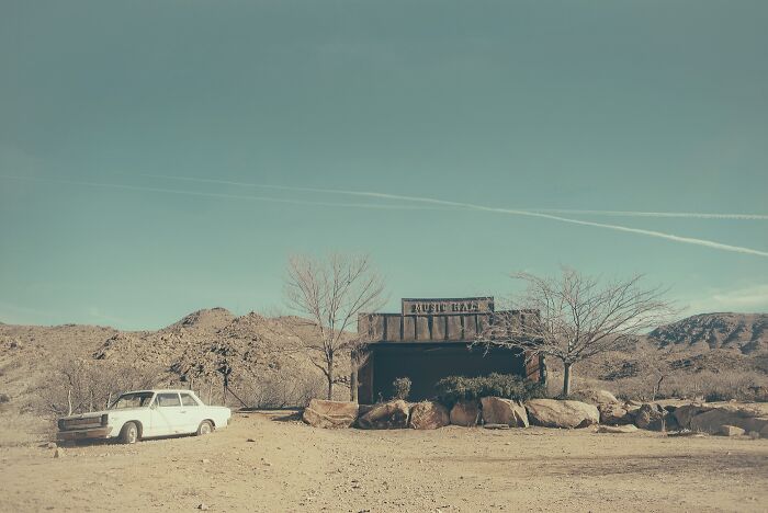 An abandoned white car sits next to a dilapidated building with 'MUSIC HALL' sign in the dusty American West. Rusty and dusty remnants.