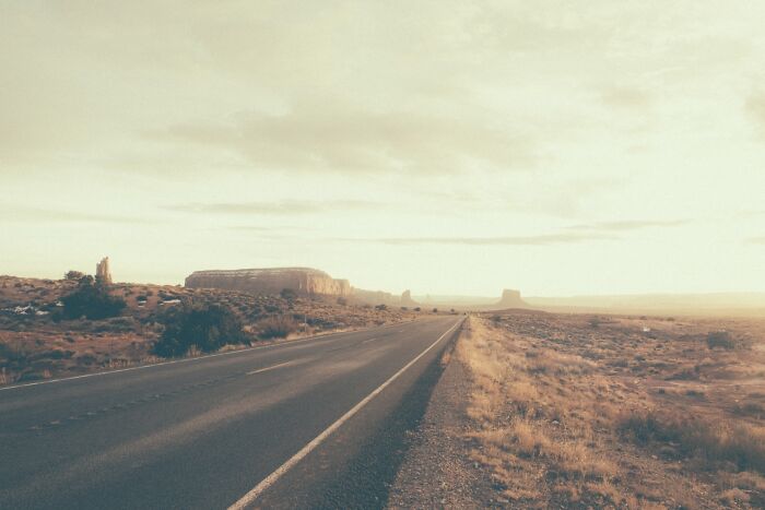 A long, empty road stretches through the American West, under a vast, pale sky. Rusty and dusty remnants.