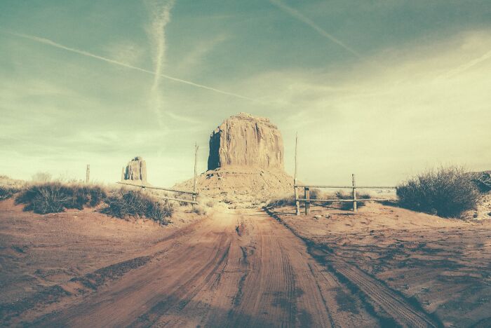 A dirt road leading towards a large rock formation in the dusty American West, with a wooden fence.