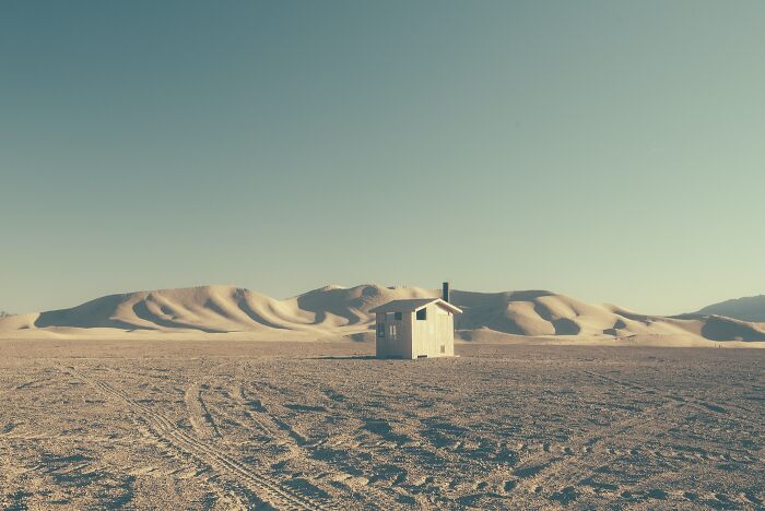 A solitary wooden shack in the dusty American West, amidst rolling sand dunes under a clear sky.