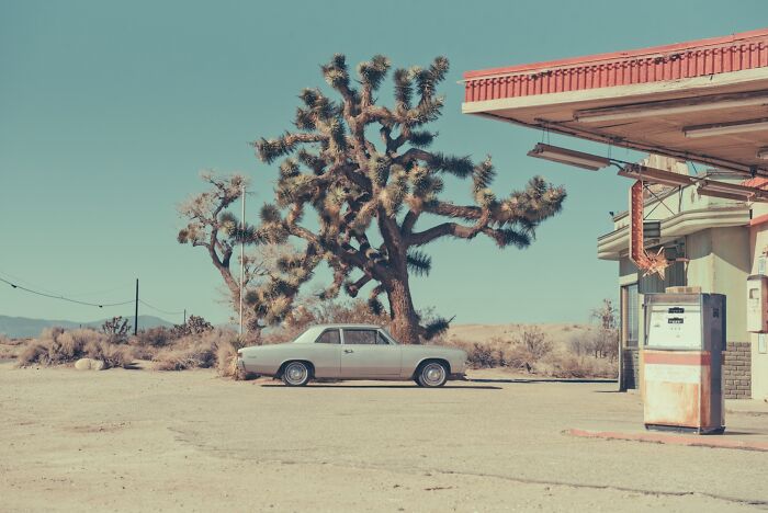 An old car parked next to a rusty gas pump and a large Joshua tree, evoking the dusty American West.