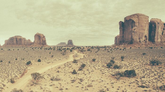 A winding dirt road through the dusty American West, with iconic rock formations under a cloudy sky.