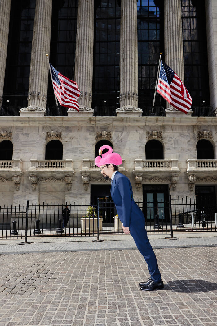 Surreal photo of man in suit wearing pink flamingo hat standing in front of large building with American flags