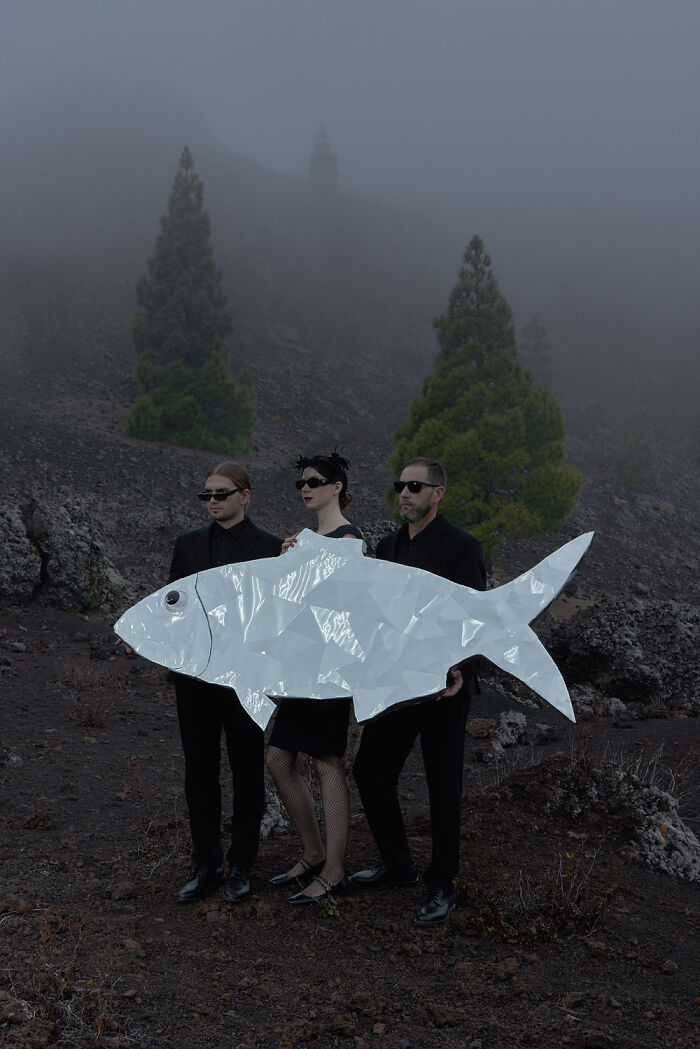 Surreal photo of three people in black holding a large silver fish-shaped object in foggy landscape