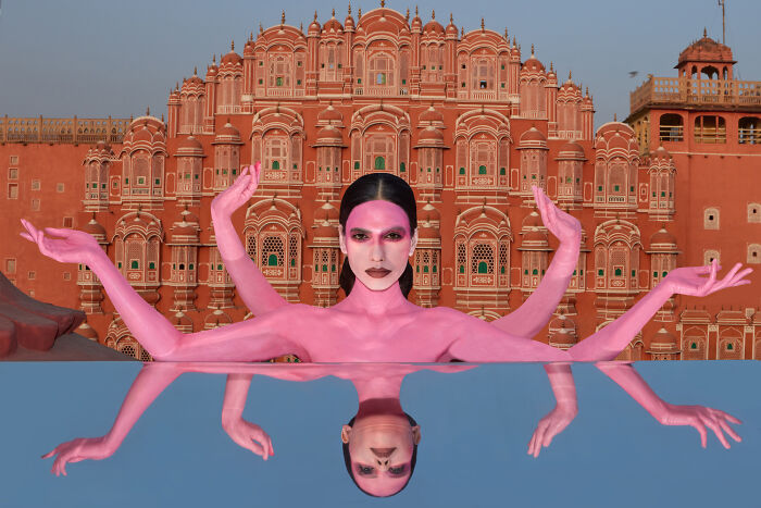 Surreal art expedition photo pink figure with multiple arms in front of ornate building with reflection