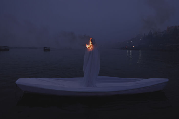 Surreal art expedition photo of figure wrapped in white on boat at night with flame