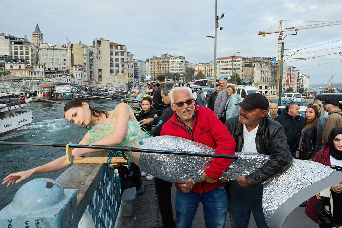 Surreal art expedition photo of model posing with giant sparkling fish sculpture on busy urban pier