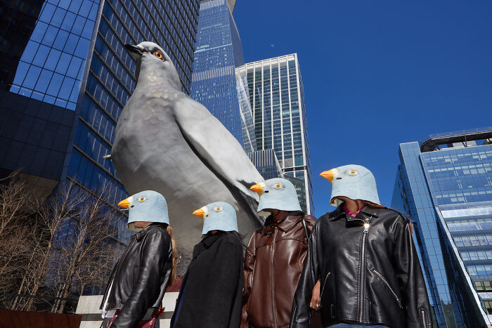 Surreal image of people wearing bird masks beside giant pigeon sculpture in city