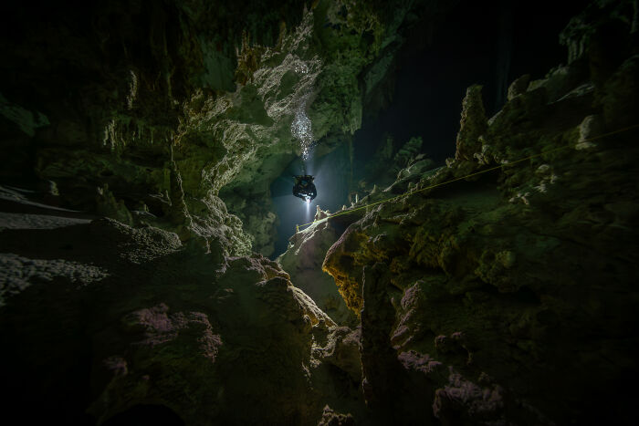 A diver descends into a dark underwater cave, illuminated by a beam of light, showcasing stunning Ocean Photographer images.