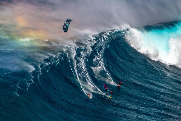 Three surfers on a huge wave with a rainbow in the background, captured in an incredible Ocean Photographer of the Year image.